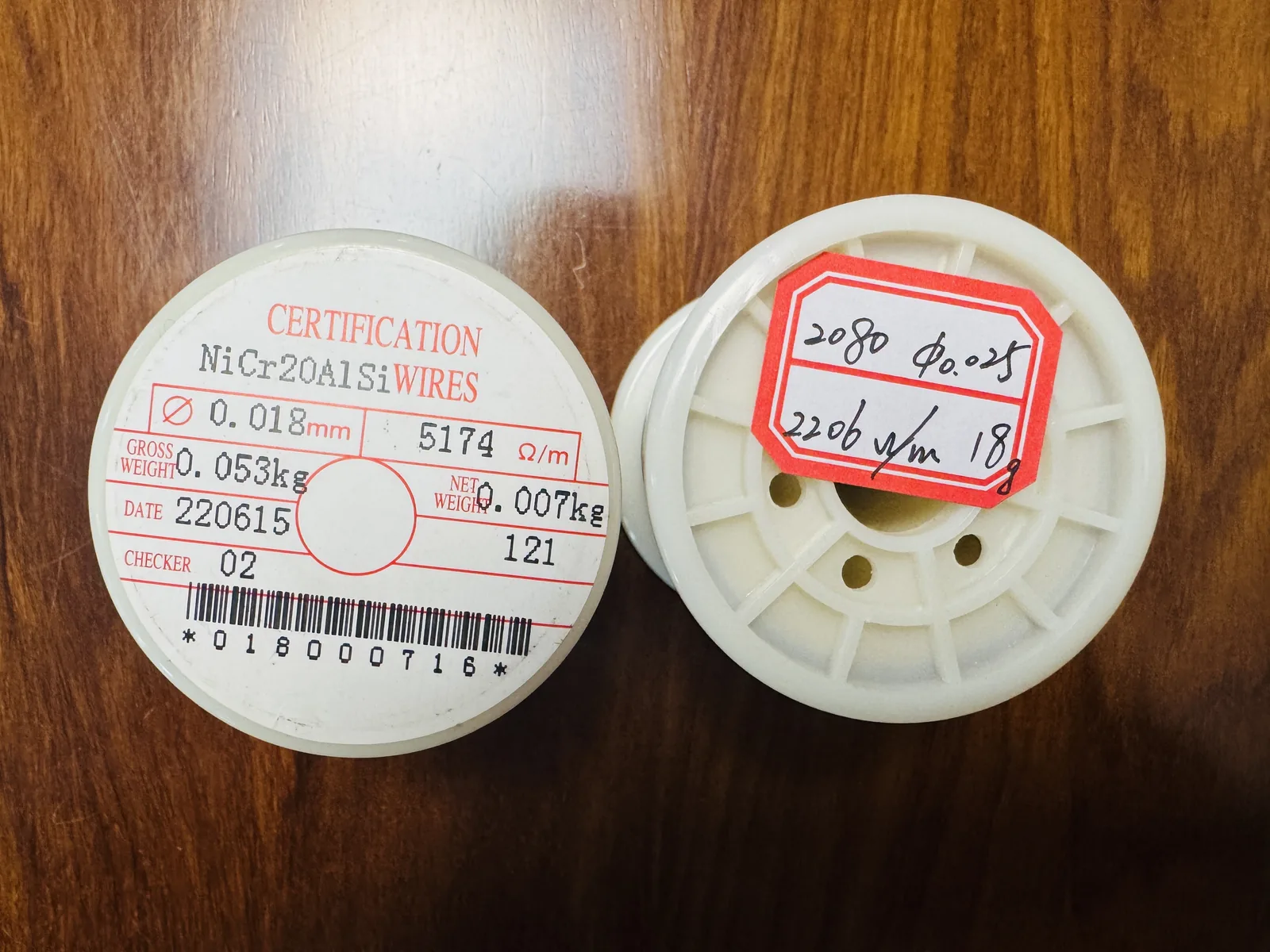 Two white-flanged spools of NiCr20AlSi wire on a wooden bench. The printed label on the left spool reads certification, NiCr20AlSi wires, 0.018 mm diameter, 5174 ohm per metre, gross weight 0.053 kg, net weight 0.007 kg, date 220615, checker 02. The red handwritten label on the right spool reads 0.025 mm and 2206 ohm per metre.