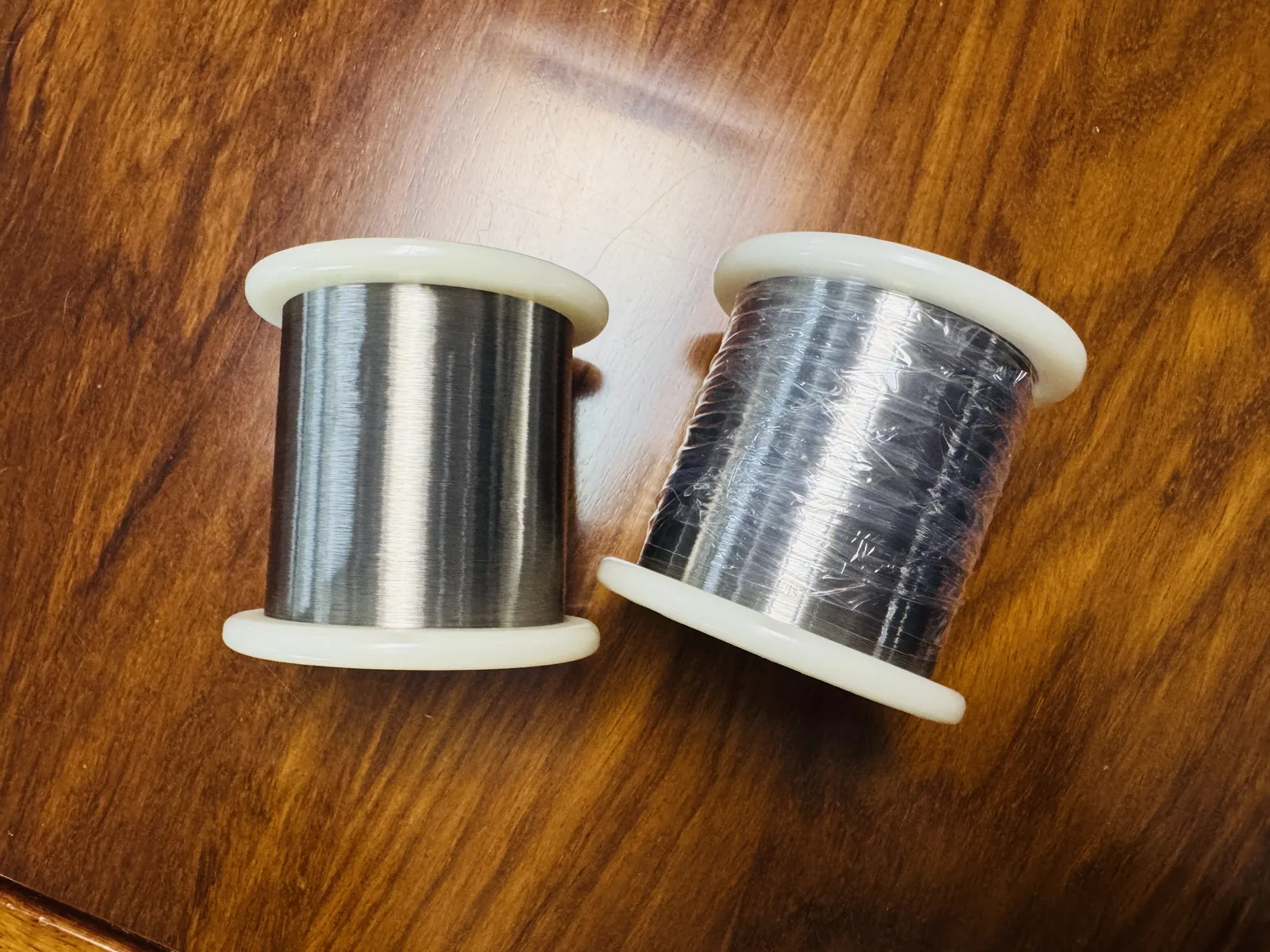 Two white-flanged spools of bright drawn alloy wire on a wooden bench, the spool on the left bare and the spool on the right still wrapped in clear film